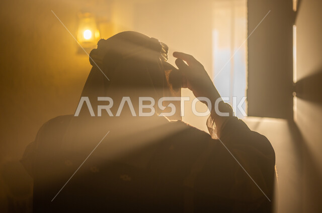 الاهتمام بالأناقة والمظهر الخارجي, مفهوم الشموخ والاعتزاز والفخر, الأعياد والمناسبات الوطنية, ذكرى تأسيس الدولة السعودية الأولى 22 فبراير, صورة مقربة من الخلف لرجل عربي خليجي سعودي يرتدي البشت والعقال يقوم بتعديل لباسه يقف بجانب النافذة, يوم بدينا 1727 م
