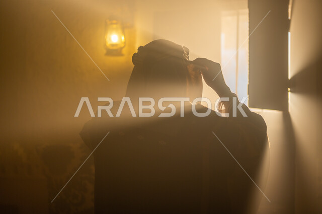 الاهتمام بالأناقة والمظهر الخارجي, مفهوم الشموخ والاعتزاز والفخر, الأعياد والمناسبات الوطنية, ذكرى تأسيس الدولة السعودية الأولى 22 فبراير, صورة مقربة من الخلف لرجل عربي خليجي سعودي يرتدي البشت والعقال يقوم بتعديل لباسه يقف بجانب النافذة, يوم بدينا 1727 م