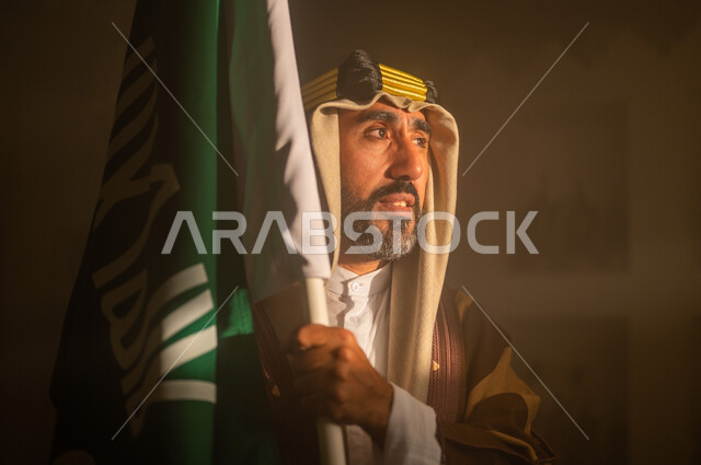 الوقوف بفخر واعتزاز, شعار الهوية الوطنية وراية الإسلام والتوحيد, ارتداء الأزياء الشعبية في ذكرى تأسيس الدولة السعودية الأولى 22 فبراير, صورة مقربة لرجل عربي خليجي سعودي يرتدي البشت والعقال يمسك علم المملكة, يوم بدينا 1727 م, مشاعر الحب والانتماء للوطن