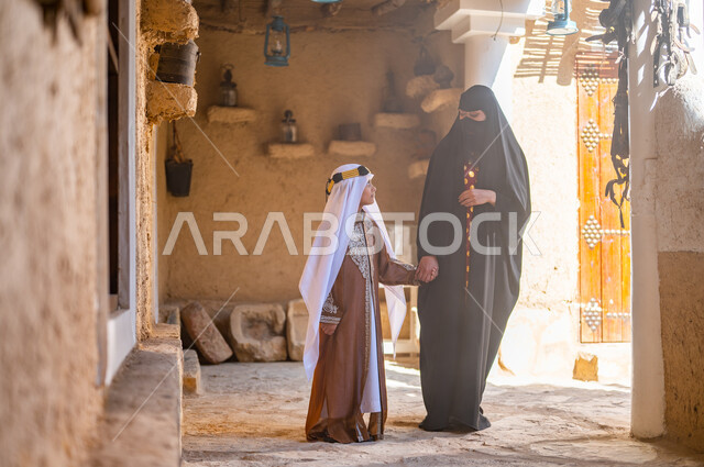تبادل الأحاديث والدردشات، ذكرى تأسيس الدولة السعودية 1727 م، تربيه الأبناء على حب الوطن، امرأة عربيه خليجيه سعوديه ترتدي البرقع والعباءه السوداء تمسك بيد ابنها وتمشي في إحدى البيوت التراثيه القديمة، ارتداء الأزياء الشعبيه التقليدية في المناسبات والأعياد الوطنية