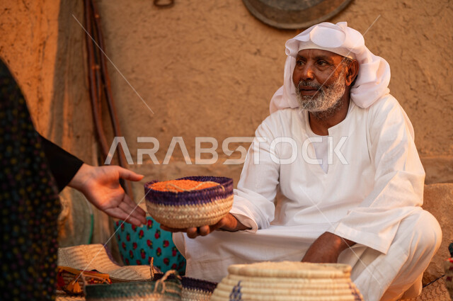 Anniversary of the founding of the first Saudi state in 1727 AD, display of national local products in baskets of handmade mats, pride in Saudi heritage, a close-up of a woman’s hand making purchases from a Saudi Arabian Gulf seller wearing traditional clothing, selling lentils and legumes on a popular rug, Badina Day, February 22