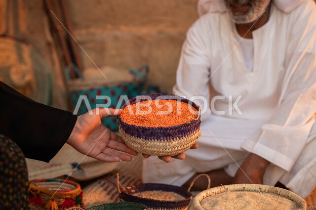 عرض المنتجات المحليه الوطنيه في سلل من الحصير اليدوي ،الفخر بالتراث السعودي ،صورة مقربة ليد امرأة تقوم بالشراء من بائع عربي خليجي سعودي يرتدي الزي التقليدي يقوم ببيع العدس و البقوليات على بساط شعبي ،يوم بدينا 22 فبراير، ذكرى تأسيس الدولة السعودية الأولى 1727 م
