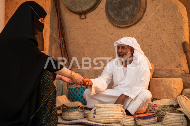 الفخر بالتراث السعودي ،صورة من الخلف لامرأة تقوم بالشراء من بائع عربي خليجي سعودي يرتدي الزي التقليدي يقوم ببيع العدس و البقوليات على بساط شعبي ،عرض المنتجات المحليه الوطنيه في سلل من الحصير اليدوي ،يوم بدينا 22 فبراير، ذكرى تأسيس الدولة السعودية الأولى 1727 م