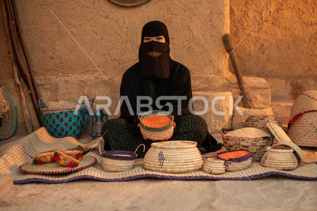 النظر إلى الكاميرا بإيماءات الفخر بالتراث السعودي ،بائعه عربيه خليجية سعودية ترتدي البرقع والعباءه تقوم ببيع العدس و البقوليات على بساط شعبي ،عرض المنتجات المحليه الوطنيه في سلل من الحصير اليدوي ، يوم بدينا 22 فبراير، ذكرى تأسيس الدولة السعودية الأولى 1727 م