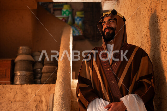 يوم بدينا 1727 م ،الوقوف بإيماءات أيدي متماسكة على درج مبني من الطين في احد البيوت التراثية، ارتداء الأزياء الشعبيه في الأعياد و المناسبات الوطنية ،صورة مقربة لرجل عربي خليجي سعودي مبتسم يرتدي الزي التقليدي في ذكرى تأسيس الدولة السعودية الاولى 22 فبراير