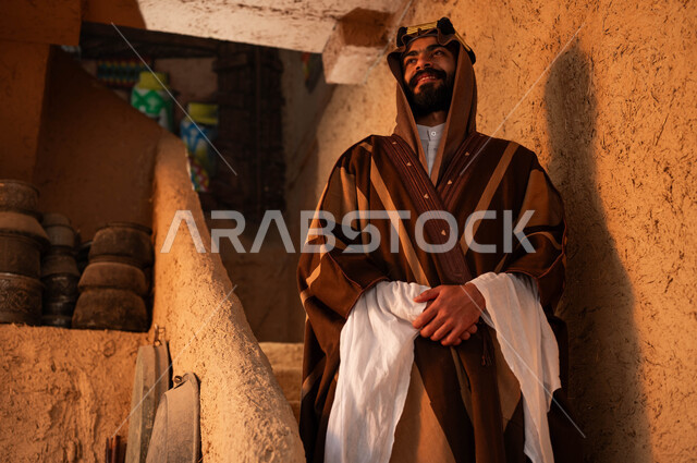 الوقوف بإيماءات أيدي متماسكة على درج مبني من الطين في احد البيوت التراثية، ارتداء الأزياء الشعبيه في الأعياد الوطنية ،رجل عربي خليجي سعودي مبتسم يرتدي الزي التقليدي في ذكرى تأسيس الدولة السعودية الاولى 22 فبراير، يوم بدينا 1727 م ،الاعتزاز بالانتماء للبلاد