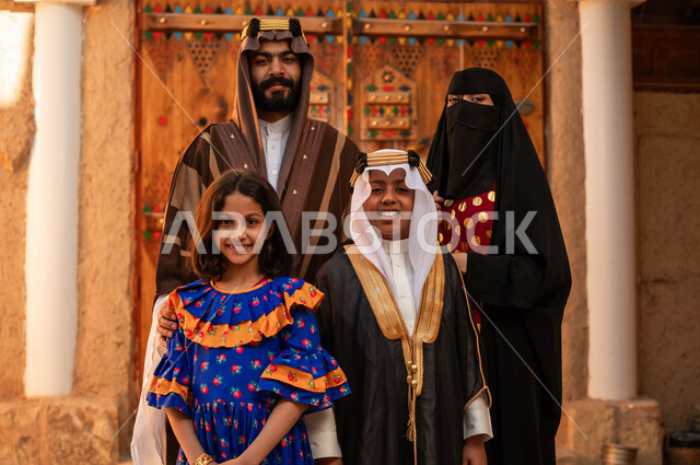 ذكرى تأسيس الدولة السعودية الأولى 1727 م، عائلة عربيه خليجية سعودية ترتدي الأزياء الشعبية في الأعياد والمناسبات الوطنية يقفون في أحد البيوت التراثية المبنية من الطين، النظر إلى الكاميرا بإيماءات الاعتزاز و الفخر بالهوية السعودية، مفهوم حب الوطن والانتماء اليه