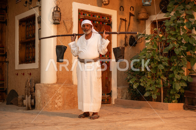 مفهوم الحياة الشعبية البدوية ،رجل مسن عربي خليجي سعودي يرتدي الزي التقليدي ويحمل على ظهره دلو شعبي من الجلد و ينظر الى الكاميرا ،سقاي الماء يقوم بالتجول في الحاره القديمه لسقي الناس من البئر ،يوم بدينا 1727 م ،ذكرى تأسيس الدولة السعودية الاولى 22 فبراير