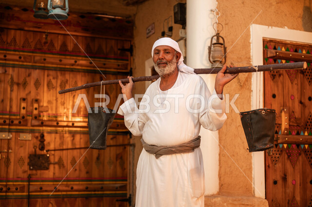 سقاي الماء يقوم بالتجول في الحاره القديمه لسقي الناس من البئر ،مفهوم الحياة الشعبية البدوية ،رجل مسن عربي خليجي سعودي يرتدي الزي التقليدي ويحمل على ظهره دلو شعبي من الجلد و ينظر الى الكاميرا ،يوم بدينا 1727 م ،ذكرى تأسيس الدولة السعودية الاولى 22 فبراير