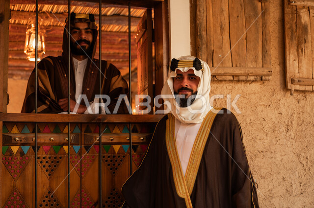 النظر الى الكاميرا بإيماءات السرور والفخر بالهوية السعودية، صورة مقربة لرجلان عربيان خليجيان سعوديان يرتديان الزي التقليدي  الشعبي للمناسبات الوطنية، ذكرى تأسيس الدولة السعودية الأولى 22 فبراير، يوم بدينا 1727 م، الوقوف في أحد المباني والبيوت الطينية التراثية
