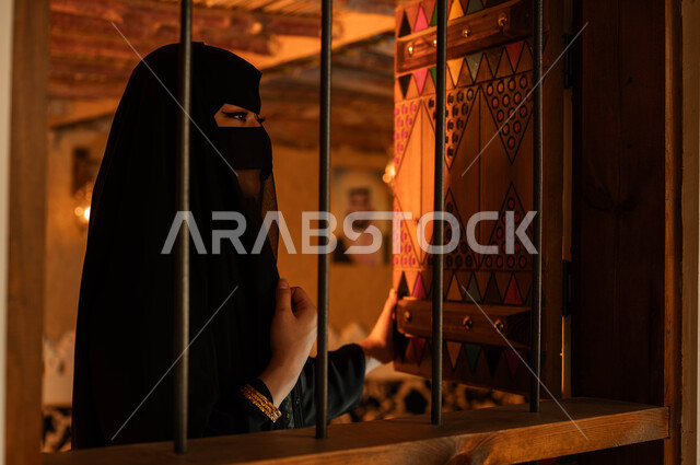 ارتداء الأزياء الشعبية التقليدية في الأعياد والمناسبات الوطنية, النظر الى شيء ،ذكرى تأسيس الدولة السعودية الأولى 22 فبراير, يوم بدينا 1727م, صورة مقربة لإمرأة عربية خليجية سعودية ترتدي العباءة والبرقع تقف بجانب النافذة داخل غرفة في أحد البيوت التراثيه