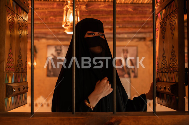 النظر الى شيء ،ذكرى تأسيس الدولة السعودية الأولى 22 فبراير, يوم بدينا 1727م, ارتداء الأزياء الشعبية التقليدية في الأعياد والمناسبات الوطنية, صورة مقربة لإمرأة عربية خليجية سعودية ترتدي العباءة والبرقع تقف بجانب النافذة داخل غرفة في أحد البيوت التراثيه