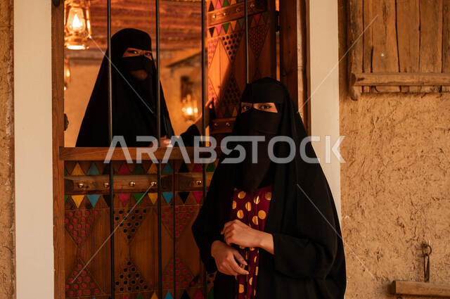 ذكرى تأسيس الدولة السعودية الأولى 22 فبراير, يوم بدينا 1727م, ارتداء الأزياء الشعبية التقليدية في الأعياد والمناسبات الوطنية, صورة مقربة لإمرأتان عربيتان خليجيتان سعوديتان ترتديان العباءة والبرقع تقفان بجانب النافذة تتحادثان, البيوت التراثيه الشعبية الطينية