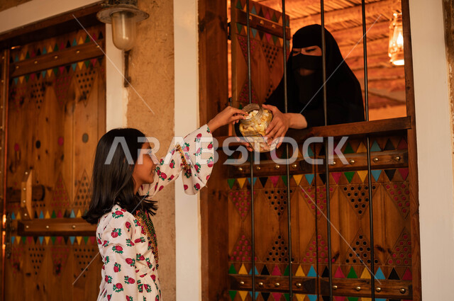 ارتداء الأزياء الشعبية التقليدية في الأعياد والمناسبات الوطنية, ايماءات السرور والفرح بالاحتفال في ذكرى تأسيس الدولة السعودية الأولى 22 فبراير, إمرأة عربية خليجية سعودية ترتدي البرقع والعباءة السوداء تقوم بتقديم الحلوى لفتاة من النافذة, يوم بدينا 1727م