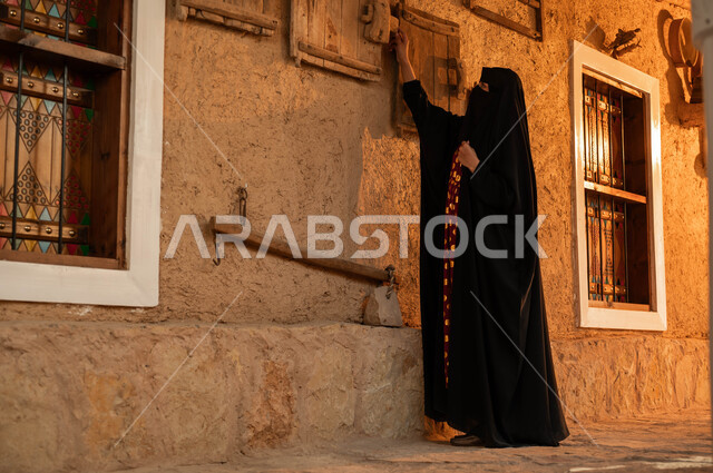 التراث السعودي العريق, ذكرى تأسيس الدولة السعودية الأولى 22 فبراير, يوم بدينا 1727م, ارتداء الأزياء الشعبية التقليدية في الأعياد والمناسبات الوطنية, إمرأة عربية خليجية سعودية ترتدي العباءة والبرقع تمسك بيدها القطع الخشبية في أحد البيوت التراثيه