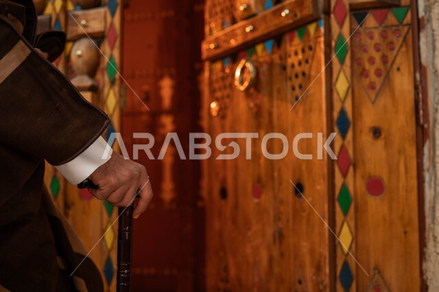 يوم بدينا 1727 م، ذكرى تأسيس الدولة السعودية الاولى 22 فبراير، الاحتفال في الأعياد والمناسبات الوطنية، مفهوم الحضارة والأصالة والعراقة، صورة مقربة ليد رجل كبير السن عربي خليجي سعودي يرتدي أزياء التأسيس يستخدم العكاز، الأبواب الخشبية المزخرفة