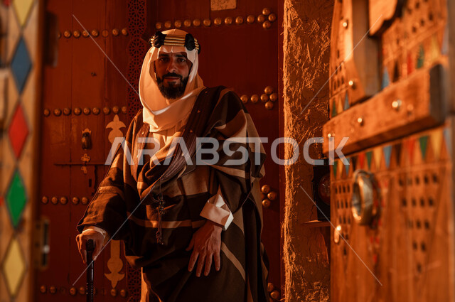 الأعياد والمناسبات الوطنية، مفهوم الحضارة والأصالة والعراقة، ذكرى تأسيس الدولة السعودية الاولى 22 فبراير، يوم بدينا 1727 م، صورة مقربة لرجل كبير السن عربي خليجي سعودي يرتدي أزياء التأسيس ينظر إلى الكاميرا يستخدم العكاز، الأبواب الخشبية المزخرفة