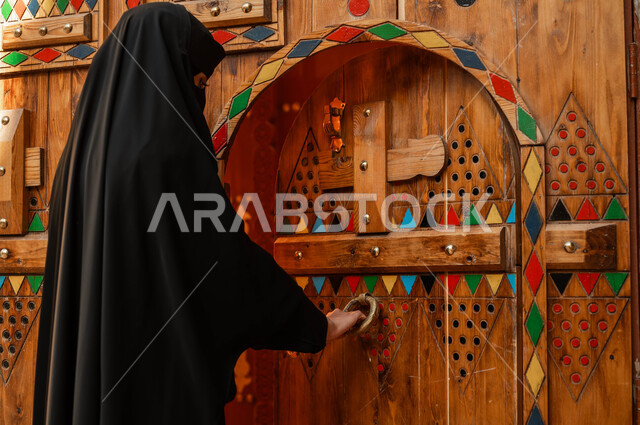 ذكرى تأسيس الدولة السعودية الاولى 22 فبراير، يوم بدينا 1727 م، ارتداء أزياء يوم التأسيس الشعبية، صورة مقربة من الخلف لامرأة عربية خليجية سعودية ترتدي العباءة السوداء والبرقع تقوم بطرق باب خشبي، نمط الحياة في البيوت الطينية التراثية