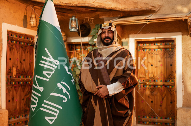 شعار الهوية الوطنية وراية الإسلام والتوحيد، الاعتزاز والفخر بإنجازات الوطن، يوم بدينا 1727 م، رجل عربي خليجي سعودي يقف بجانب علم التأسيس في بيت طيني تراثي, ذكرى تأسيس الدولة السعودية الأولى 22 فبراير، المناسبات والأعياد الوطنية