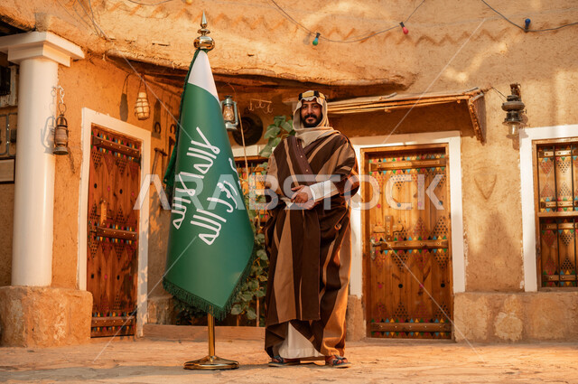 شعار الهوية الوطنية وراية الإسلام والتوحيد، الاعتزاز والفخر بإنجازات الوطن، يوم بدينا 1727 م، رجل عربي خليجي سعودي يقف بجانب علم التأسيس في بيت طيني تراثي, ذكرى تأسيس الدولة السعودية الأولى 22 فبراير، المناسبات والأعياد الوطنية, صورة الجسم بالطول الكامل