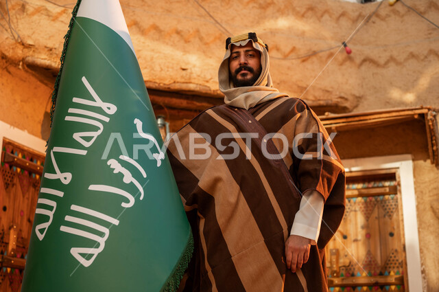 شعار الهوية الوطنية وراية الإسلام والتوحيد، الاعتزاز والفخر بإنجازات الوطن، يوم بدينا 1727 م، رجل عربي خليجي سعودي يقف بجانب علم التأسيس في بيت طيني تراثي, ذكرى تأسيس الدولة السعودية الأولى 22 فبراير، المناسبات والأعياد الوطنية