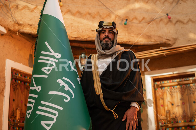 الفخر والاعتزاز بإنجازات الوطن، شعار الهوية الوطنية وراية الإسلام والتوحيد، يوم بدينا 1727 م، رجل عربي خليجي سعودي يقف بجانب علم التأسيس في بيت طيني تراثي, ذكرى تأسيس الدولة السعودية الأولى 22 فبراير، المناسبات والأعياد الوطنية