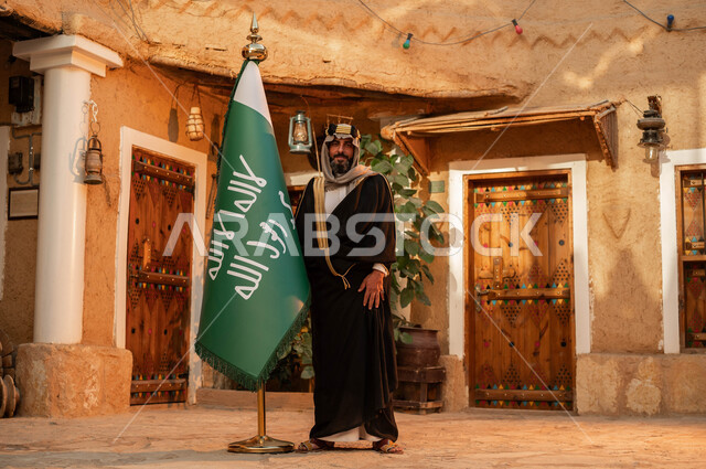 الاعتزاز والفخر بإنجازات الوطن، شعار الهوية الوطنية وراية الإسلام والتوحيد، يوم بدينا 1727 م، رجل عربي خليجي سعودي يقف بجانب علم التأسيس في بيت طيني تراثي, ذكرى تأسيس الدولة السعودية الأولى 22 فبراير، المناسبات والأعياد الوطنية, صورة الجسم بالطول الكامل