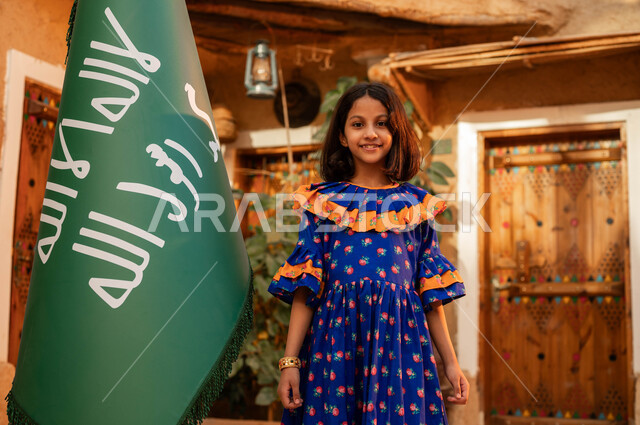 المحافظة على التراث السعودي، الوقوف بإيماءات الفخر والاعتزاز، يوم بدينا 1727 م، ذكرى تأسيس الدولة السعودية الاولى 22 فبراير، فتاة عربيه خليجية سعودية مبتسمة ترتدي الأزياء الشعبية تقف بجانب علم التأسيس، المناسبات والأعياد الوطنية، تربيه الأبناء على حب الوطن