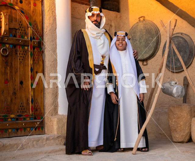 Standing with gestures of pride and pride, Day of Dina 1727 AD, the anniversary of the founding of the first Saudi state, February 22, a Saudi Gulf Arab man wearing Founding Day costumes standing with his son in front of old heritage houses looking at the camera, national occasions and holidays