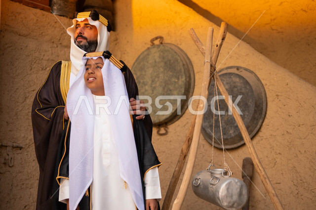 الوقوف بإيماءات الفخر والاعتزاز، يوم بدينا 1727 م، ذكرى تأسيس الدولة السعودية الاولى 22 فبراير، رجل عربي سعودي خليجي يرتدي أزياء يوم التأسيس يقف مع ولده أمام البيوت التراثية القديمة، المناسبات والأعياد الوطنية