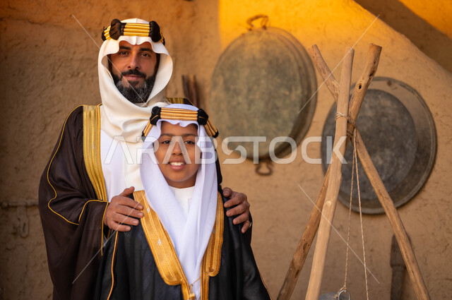 النظر إلى الكاميرا بإيماءات الفخر والاعتزاز، يوم بدينا 1727 م، ذكرى تأسيس الدولة السعودية الاولى 22 فبراير، رجل عربي سعودي خليجي يرتدي زي يوم التأسيس يقف مع ولده أمام البيوت التراثية القديمة، المناسبات والأعياد الوطنية