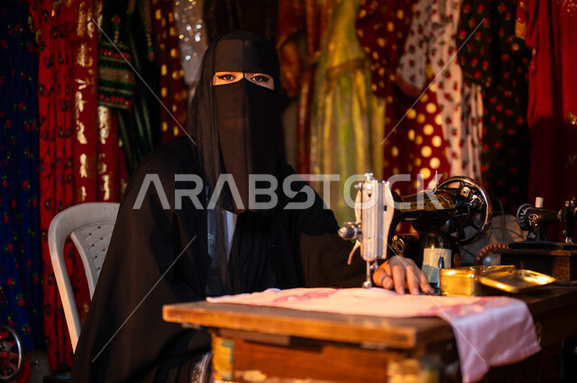 يوم بدينا 1727 م، حياكة وخياطة القماش على الماكينة اليدوية، تصميم الأزياء الشعبية التراثية، حرف وأعمال ومهن تقليدية نسائية, خياطة عربية سعودية خليجية ترتدي العباءة السوداء والبرقع تنظر إلى الكاميرا، ذكرى تأسيس الدوله السعوديه الاولى 22 فبراير