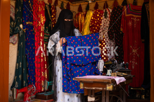 المحلات التجارية التراثية، حرف ومهن تقليدية نسائية, امرأة عربية سعودية خليجية ترتدي العباءة والبرقع تقوم بتصميم وتجهيز الأزياء الشعبية التراثية للمناسبات الوطنية، خياطة القماش على الماكينة اليدوية، ذكرى تأسيس الدوله السعوديه الاولى 22 فبراير، يوم بدينا 1727 م