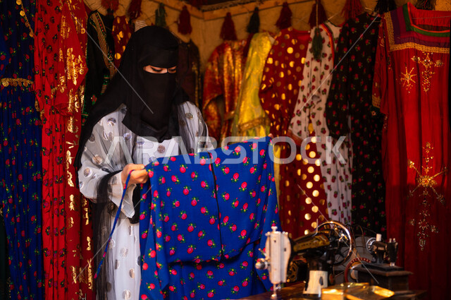 المحلات التجارية التراثية، حرف ومهن تقليدية نسائية, امرأة عربية سعودية خليجية ترتدي العباءة والبرقع تقوم بتصميم وتجهيز الأزياء الشعبية التراثية للمناسبات الوطنية، خياطة القماش على الماكينة اليدوية، ذكرى تأسيس الدوله السعوديه الاولى 22 فبراير، يوم بدينا 1727 م