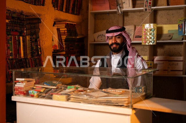 ذكرى تأسيس الدولة السعودية الأولى 22 فبراير، يوم بدينا 1727 م، المكتبات القديمة في الأحياء التراثية الطينية، النظر إلى الكاميرا بإيماءات السعادة, أماكن بيع الكتب التاريخية والمستلزمات المكتبية, شاب عربي خليجي سعودي مبتسم يرتدي الزي التقليدي يجلس داخل مكتبته