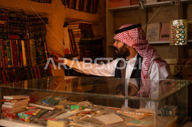 حب الثقافة والطلاع، دكان قديم لبيع الكتب والمستلزمات المكتبية, المكتبات القديمة في الأحياء التراثية الطينية، يوم بدينا 1727 م، ذكرى تأسيس الدولة السعودية الأولى 22 فبراير، شاب عربي خليجي سعودي يرتدي الزي التقليدي يقوم باختيار كتاب من المكتبة