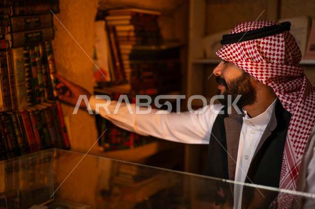 دكان قديم لبيع الكتب والمستلزمات المكتبية, ممارسة هواية القراءة, المكتبات القديمة في الأحياء التراثية الطينية، يوم بدينا 1727 م، ذكرى تأسيس الدولة السعودية الأولى 22 فبراير، صورة مقربة لشاب عربي خليجي سعودي يرتدي الزي التقليدي يقوم باختيار كتاب من المكتبة