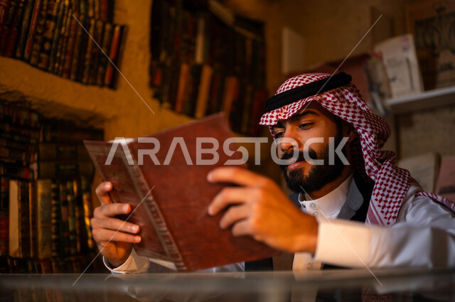 دكان قديم لبيع الكتب والمستلزمات المكتبية, ممارسة هواية المطالعة, المكتبات القديمة في الأحياء التراثية الطينية، يوم بدينا 1727 م، ذكرى تأسيس الدولة السعودية الأولى 22 فبراير، صورة مقربة لشاب عربي خليجي سعودي يرتدي الزي التقليدي يقوم بقراءة كتاب بإيماءات التركيز