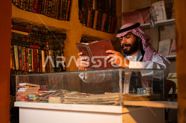 حب وشغف الثقافة والاطلاع, أماكن بيع الكتب والمستلزمات المكتبية, المكتبات القديمة في الأحياء التراثية الطينية، يوم بدينا 1727 م، ذكرى تأسيس الدولة السعودية الأولى 22 فبراير، شاب عربي خليجي سعودي يرتدي الزي التقليدي يقوم بقراءة كتاب داخل دكانه