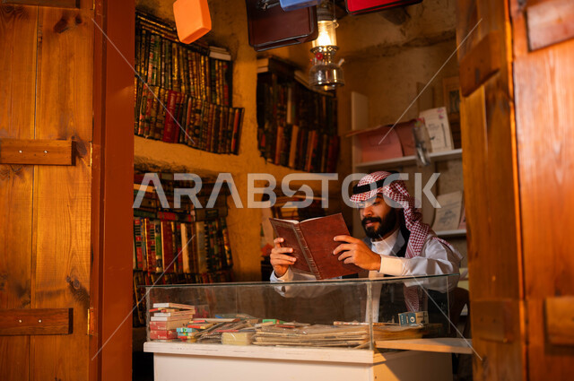أماكن بيع الكتب والمستلزمات المكتبية, المكتبات القديمة في الأحياء التراثية الطينية، يوم بدينا 1727 م، ذكرى تأسيس الدولة السعودية الأولى 22 فبراير، حب وشغف الثقافة والاطلاع, شاب عربي خليجي سعودي يرتدي الزي التقليدي يقوم بقراءة كتاب داخل دكانه