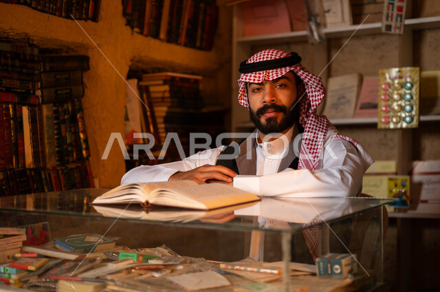 النظر إلى الكاميرا بإيماءات الثقة بالنفس, أماكن بيع الكتب والمستلزمات المكتبية, المكتبات القديمة في الأحياء التراثية الطينية، يوم بدينا 1727 م، ذكرى تأسيس الدولة السعودية الأولى 22 فبراير، شاب عربي خليجي سعودي يرتدي الزي التقليدي يجلس في دكانه