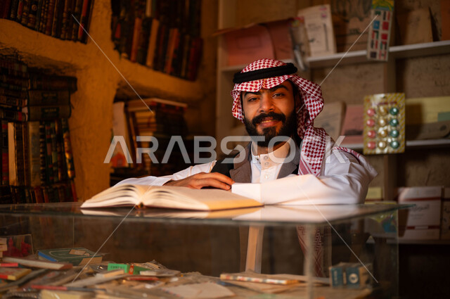 النظر إلى الكاميرا بإيماءات السرور, أماكن بيع الكتب والمستلزمات المكتبية, المكتبات القديمة في الأحياء التراثية الطينية، يوم بدينا 1727 م، ذكرى تأسيس الدولة السعودية الأولى 22 فبراير، شاب عربي خليجي سعودي مبتسم يرتدي الزي التقليدي يقوم بقراءة كتاب