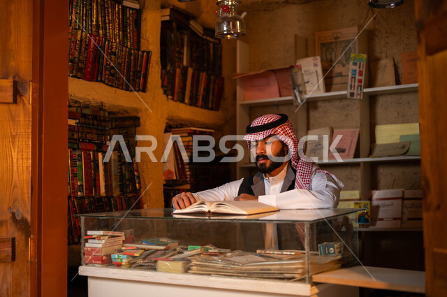 أماكن بيع الكتب والمستلزمات المكتبية, المكتبات القديمة في الأحياء التراثية الطينية، يوم بدينا 1727 م، ذكرى تأسيس الدولة السعودية الأولى 22 فبراير، حب وشغف الثقافة والاطلاع, شاب عربي خليجي سعودي يرتدي الزي التقليدي يقوم بقراءة كتاب داخل دكانه