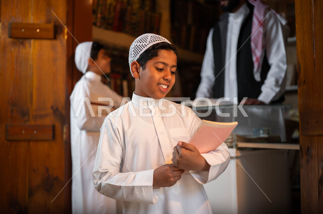 شغف الثقافة والاطلاع, فتى عربي خليجي سعودي يرتدي الثوب التقليدي والقبعة البيضاء يحمل بيده كتاب بإيماءات السعادة، مكتبة لبيع الكتب والمستلزمات المكتبية، يوم بدينا 1727 م، المكتبات القديمة في الأحياء التراثية الطينية، ذكرى تأسيس الدولة السعودية الأولى 22 فبراير