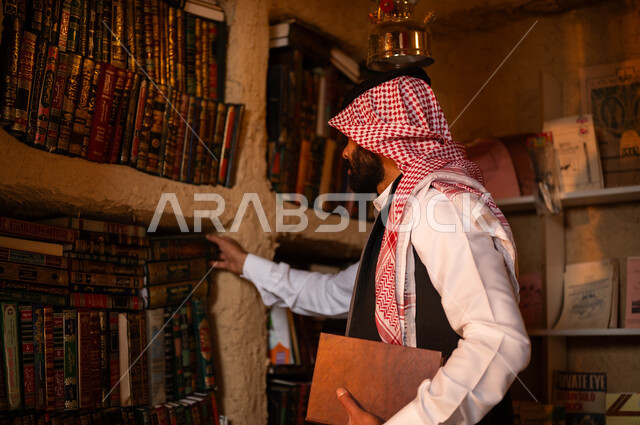 An old shop that sells books and office supplies, practicing the hobby of reading, old libraries in the mud heritage neighborhoods, Day of Dina 1727 AD, the anniversary of the founding of the first Saudi state, February 22, a young Saudi Gulf Arab man wearing traditional clothing is choosing a book from the library