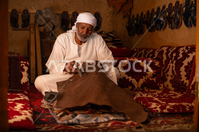 ذكرى تأسيس الدولة السعودية الأولى 22 فبراير, متجر اصلاح الأحذية, إحياء التراث الخليجي, اسكافي كبير بالسن عربي خليجي سعودي يرتدي الزي التقليدي يقوم بقص قطعة الجلد، الحرف و المهن اليدوية التقليدية القديمة, يوم بدينا 1727 م، صناعات و أعمال شعبية