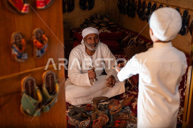 استخدام الأدوات المناسبة في تصليح الأحذية, مهن وحرف يدوية شعبية, صورة من الخلف لولد عربي خليجي سعودي يرتدي الثوب التقليدي والطاقية البيضاء يقوم بإعطاء الحذاء لصاحب الدكانة, يوم بدينا 1727م, ذكرى تأسيس الدولة السعودية الأولى في 22 فبراير, صناعات تقليدية قديمة