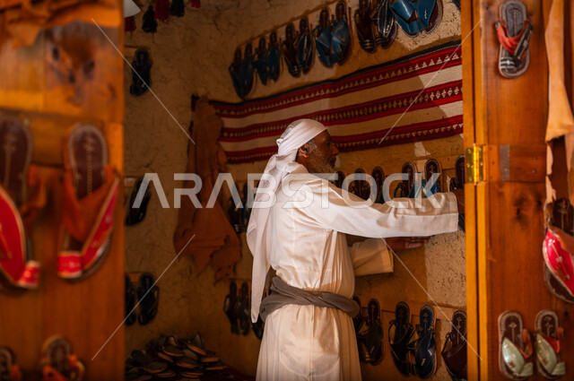 Reviving the traditional Gulf heritage, traditional works, professions and crafts, a shoe repair and sale shop, Day of Our Day 1727 AD, the anniversary of the founding of the first Saudi state, February 22, a picture from the side of an old, smiling Saudi Gulf Arab man wearing traditional clothing, displaying shoes in his shop