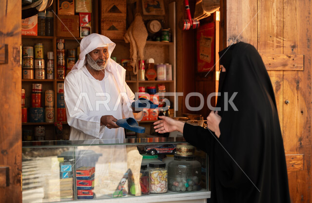 مهن تقليدية شعبية, الأسواق القديمة التراثية, محل تجاري لبيع الأغراض والمستلزمات الضرورية, صورة من الخلف لإمرأة عربية خليجية سعودية ترتدي العباءة السوداء والبرقع تقوم بشراء الشبشب من الدكان, يوم بدينا 1727م, ذكرى تأسيس الدولة السعودية الأولى في 22 فبراير