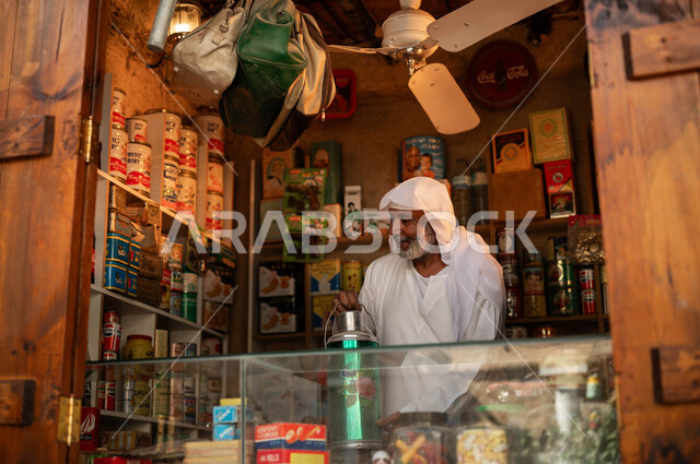 ذكرى تأسيس الدولة السعودية الأولى في 22 فبراير, المحلات الشعبية القديمة, رجل كبير بالسن عربي خليجي سعودي مبتسم يرتدي الزي التقليدي يحمل حافظة المشروبات الساخنة, أماكن بيع المستلزمات المنزلية في الأسواق التراثية السعودية, يوم بدينا 1727م